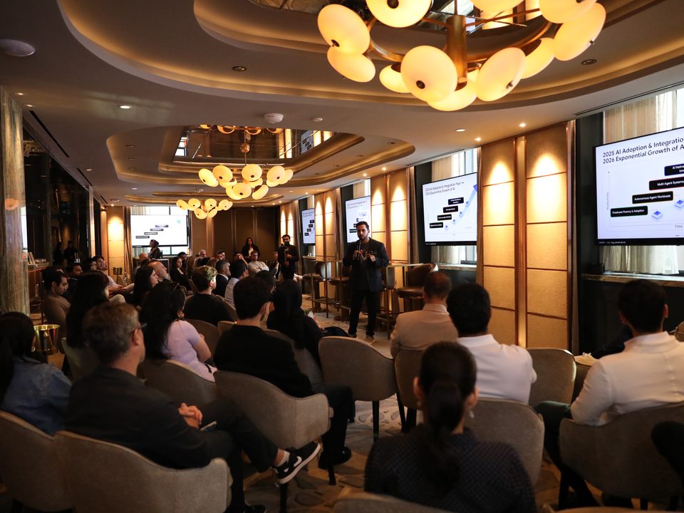 A full room at The Agentic Boardroom, with a Zero to Agent speaker standing by the screens under chandeliers.